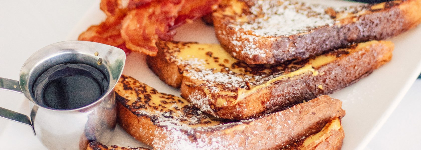 French toast sprnkled with powdered sugar on a plate with bacon and a small cup of syrup.