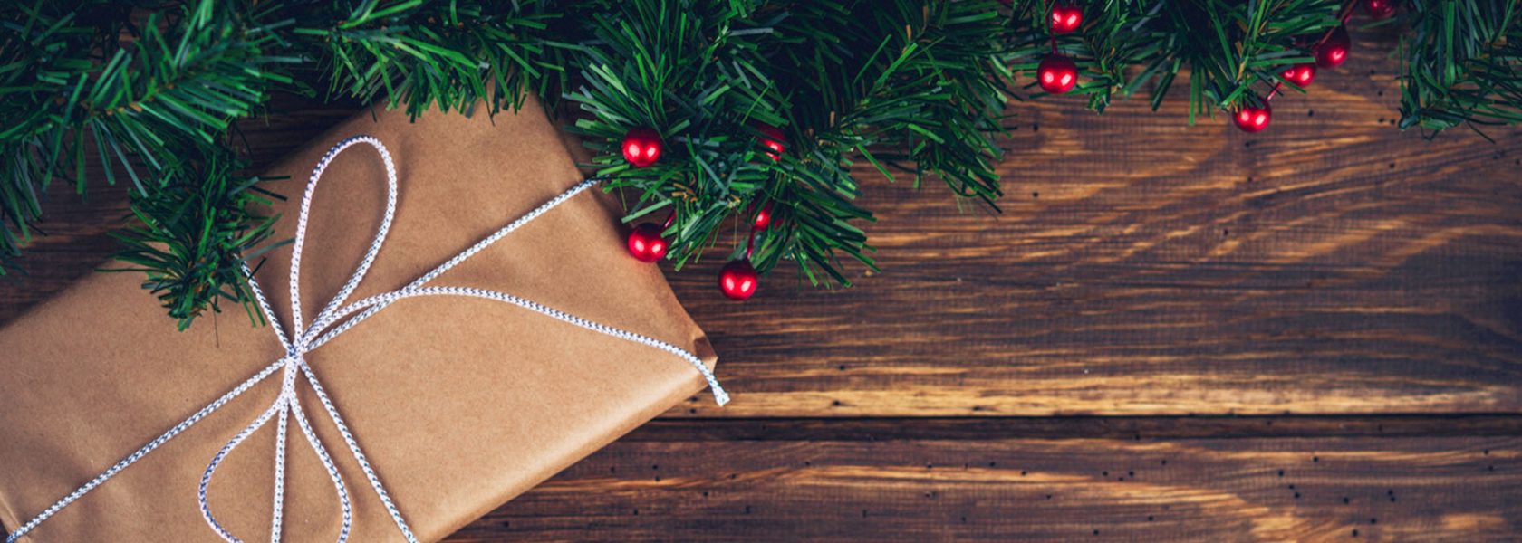 A present wrapped in brown paper and a silver bow tied around it in front of a wood detailed background and green holiday garland draped over the top of them.