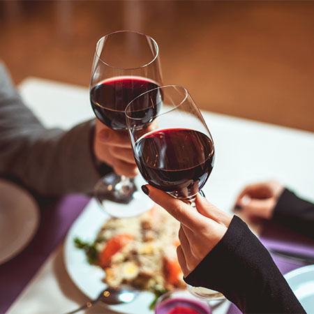 Two people clinking their full wine glasses together over dinner.