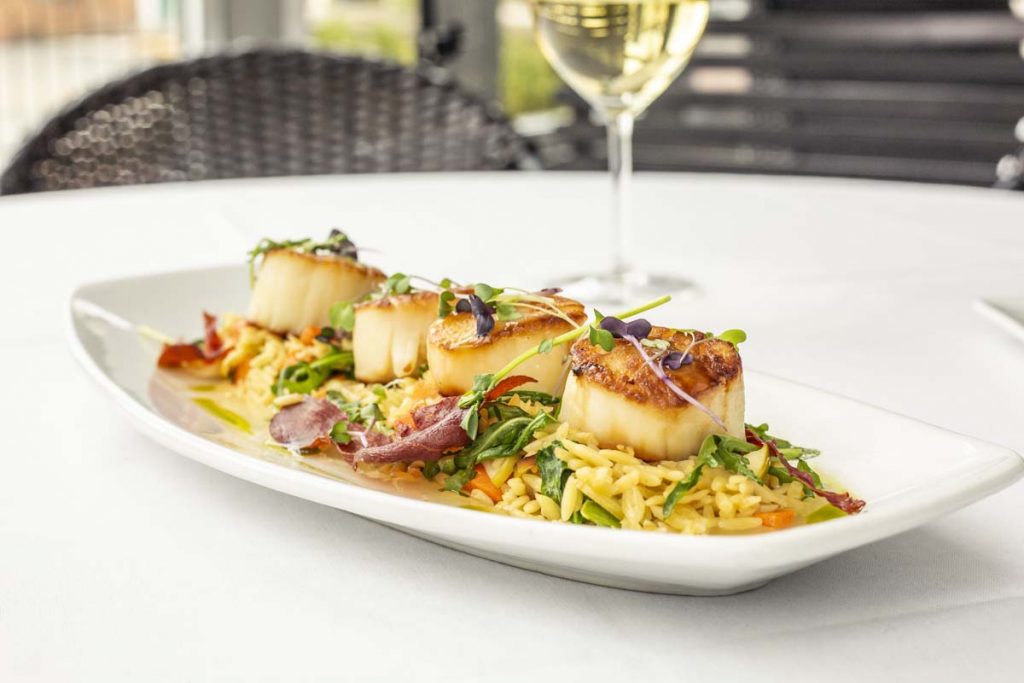 Tucci’s jumbo pan-seared scallops on bed of orzo pasta with white wine in background