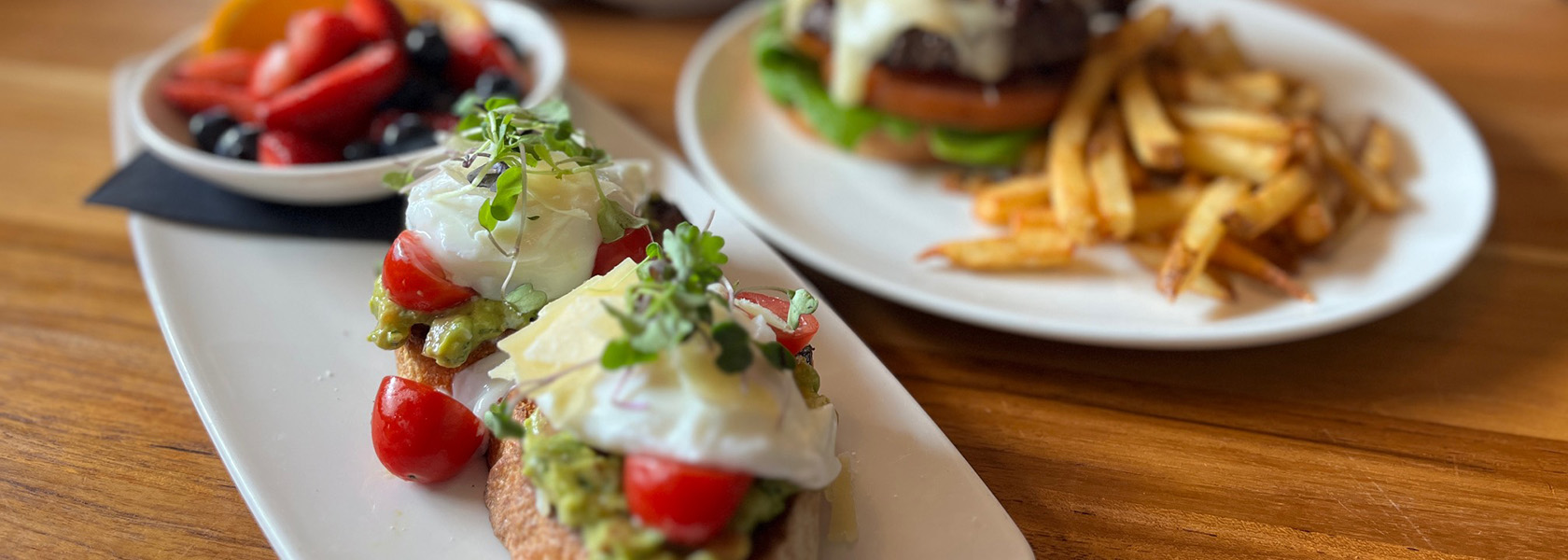 Plated avocado toast with dish of fresh fruit and burger with fries in background