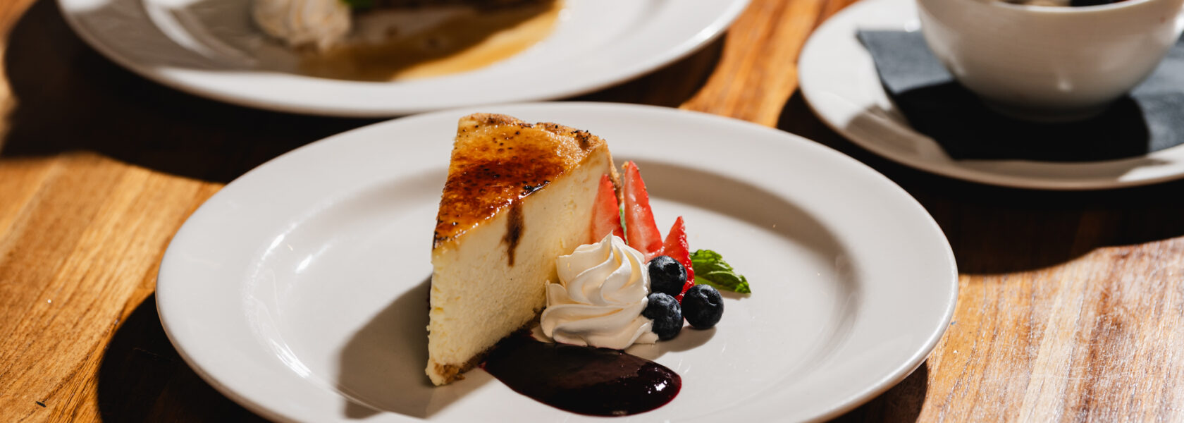 A slice of vanilla brulee cheesecake with whipped cream, berries and berry sauce on a white plate, with a slice of carrot cake behind it